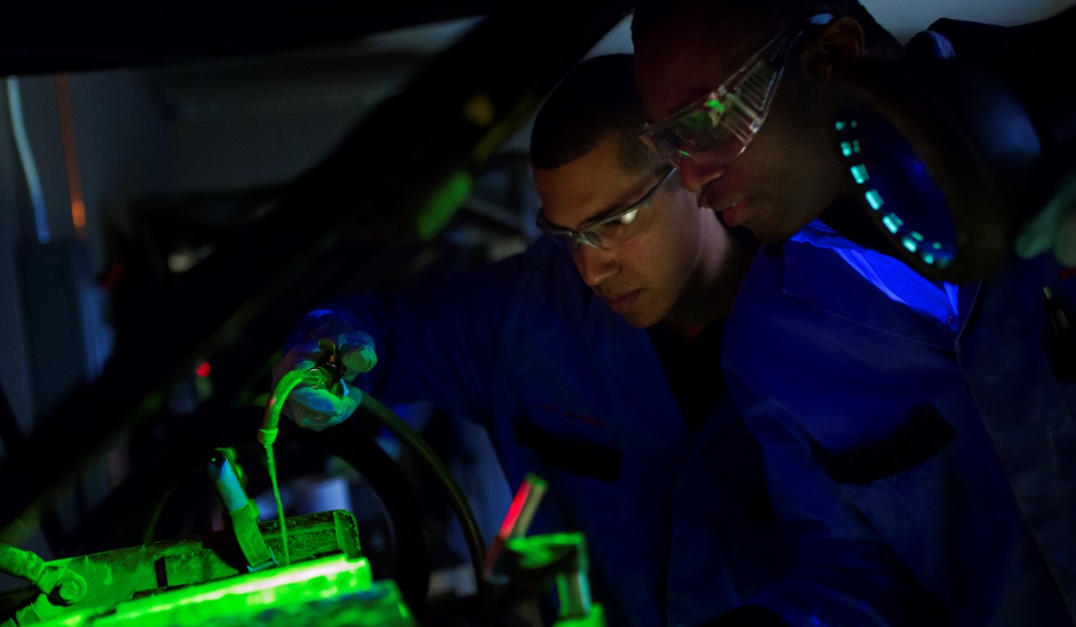 Two technicians examining a vehicle's battery under green light while wearing safety goggles and blue work uniforms.