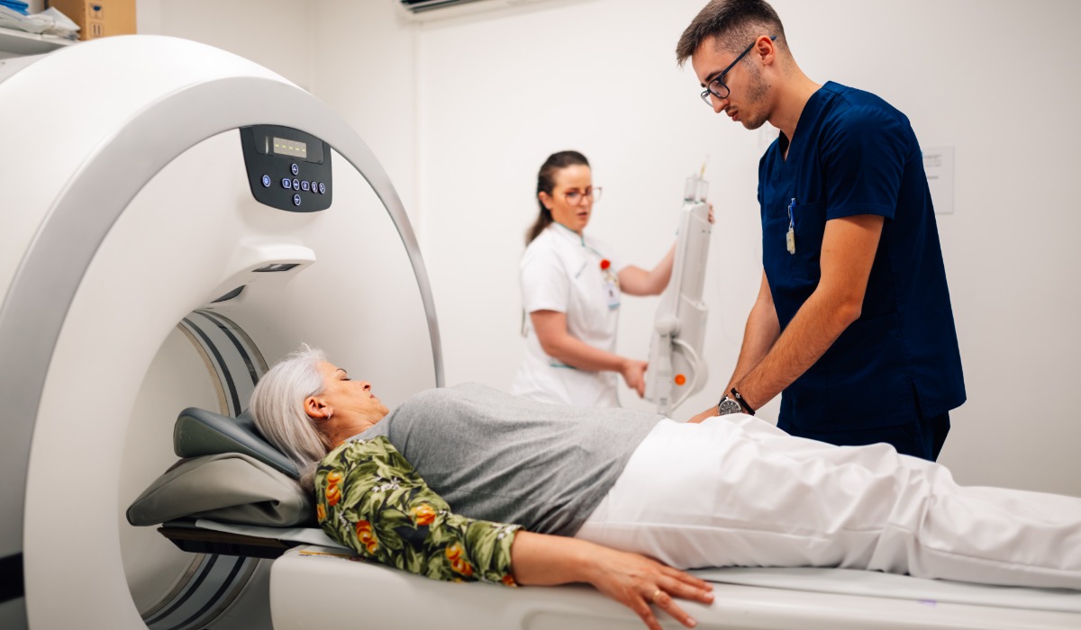 An older woman lies on a medical scanner table preparing for an imaging procedure, while a healthcare professional in blue scrubs adjusts her position and another staff member in a white uniform operates equipment in the background.