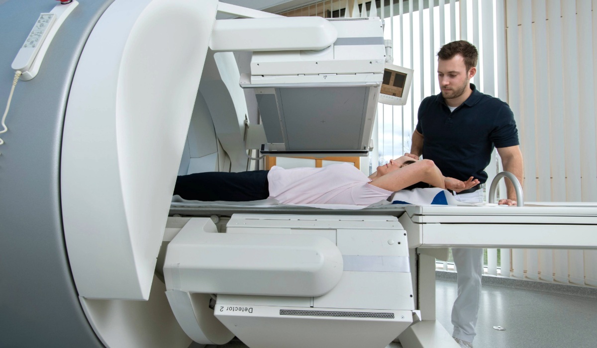 A patient lies on a scanning table beneath a large nuclear medicine imaging machine while a technician stands beside them, adjusting their head position in a clinical setting.
