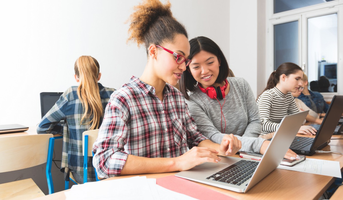 Two diverse female students collaborate on a laptop while seated at a desk in a classroom, with other students working in the background.