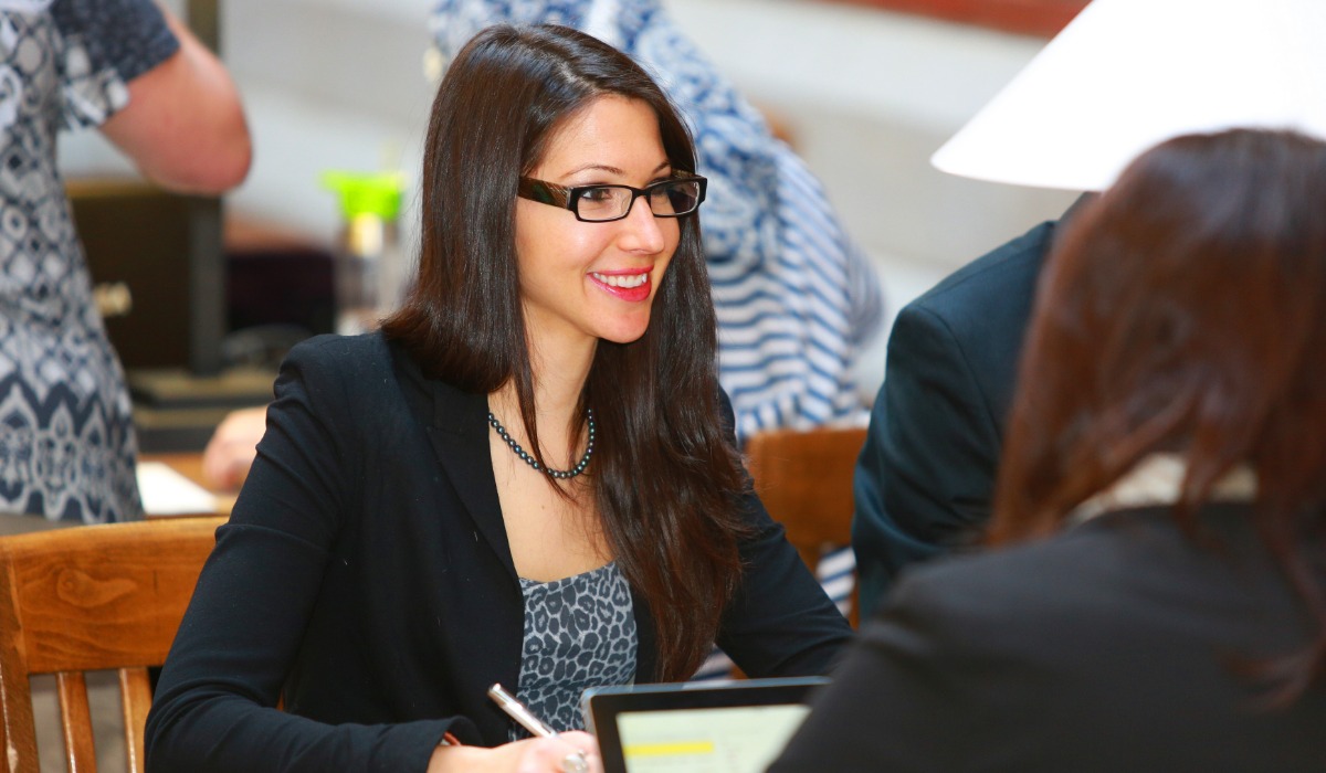 A woman in a professional outfit with glasses smiles while engaged in a conversation at a table.