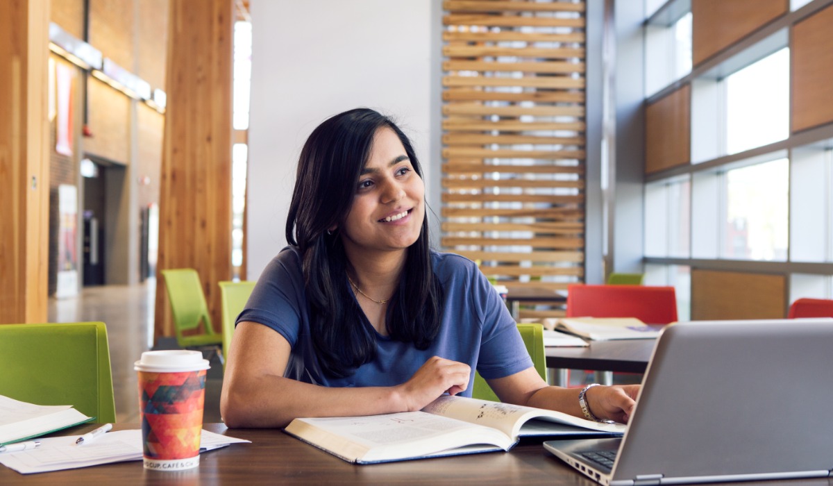 A student engaged in study with a laptop, books, and a coffee cup in a bright, modern study space.