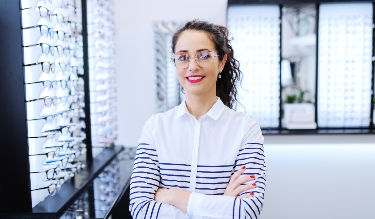 A smiling female optometric assistant stands confidently in an eyewear store, surrounded by various eyeglasses displays.