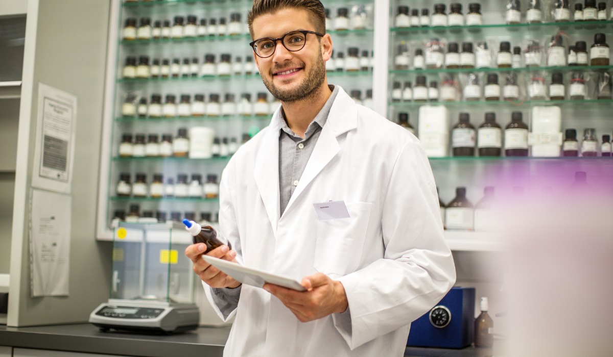 A smiling male pharmacy assistant in a lab coat holds a tablet and a bottle in a pharmacy filled with various medicinal products.