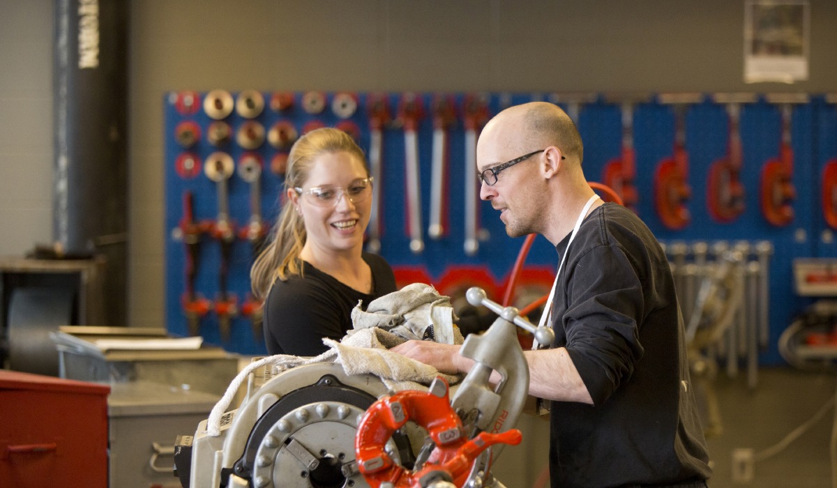 Two apprentices wearing safety glasses work together on a plumbing assembly in a workshop, with tools and equipment organized on a wall in the background.