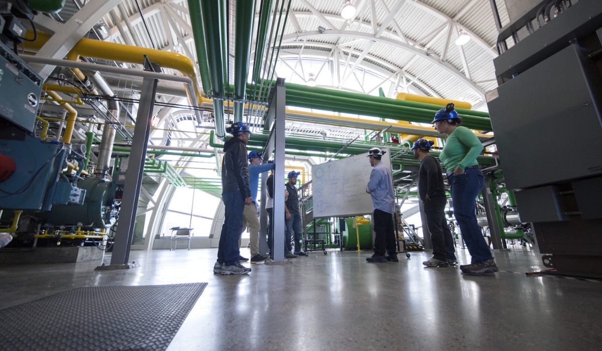 A group of students wearing safety helmets and protective gear stand around a whiteboard discussing plans inside an industrial facility with large pipes, machinery, and metal structures.