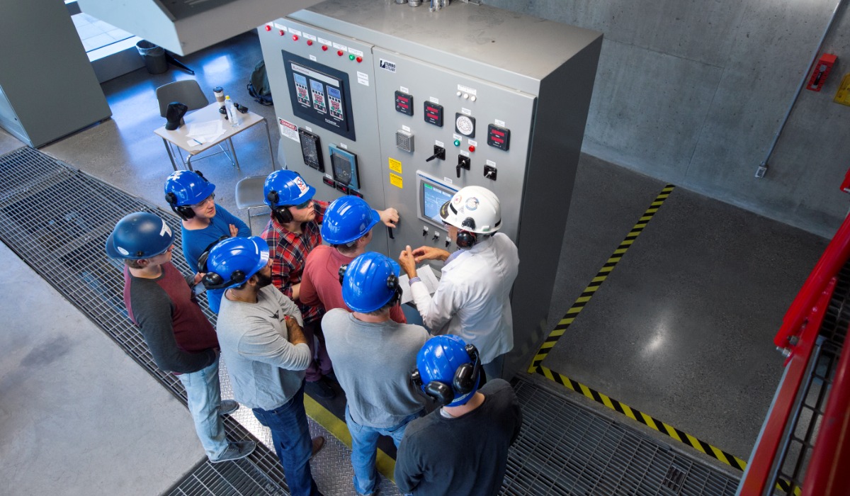 Group of workers in blue hard hats gathered around a control panel for a training session.