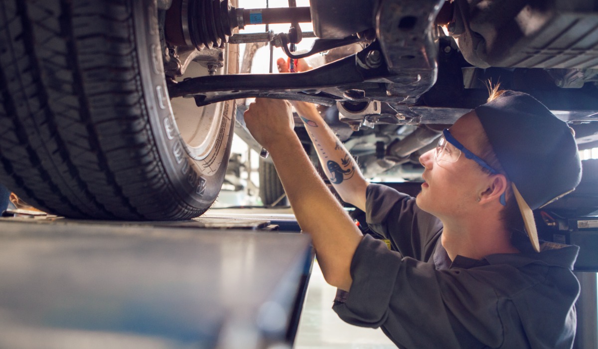 A mechanic wearing safety glasses and a backward cap works underneath a raised vehicle, using a tool to adjust components near the suspension and wheel area.