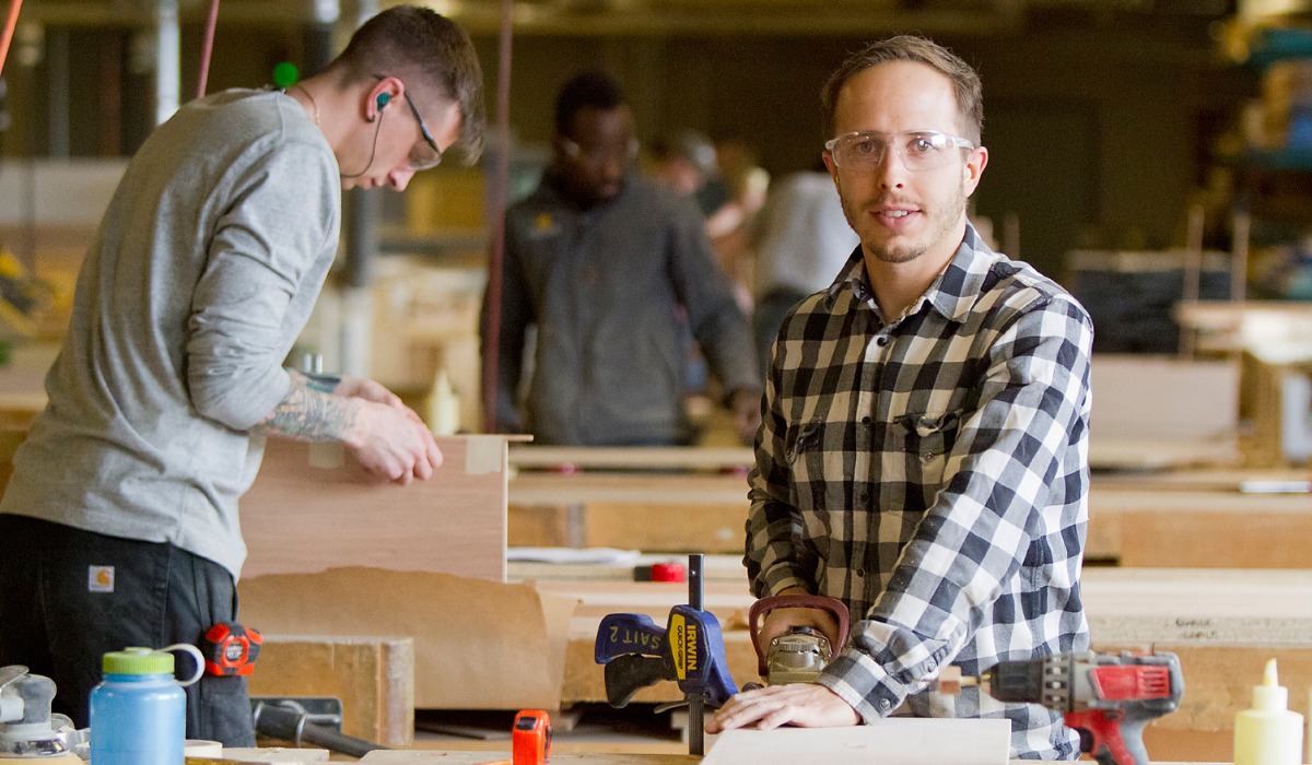A woodworking workshop scene featuring a focused carpenter in a plaid shirt with safety glasses, alongside another worker operating tools.