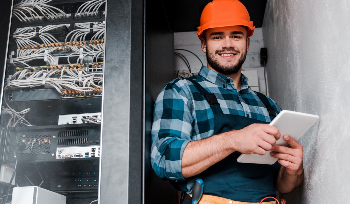 A smiling electrician wearing a hard hat and work overalls holds a tablet while standing next to a server rack filled with cables and network equipment.