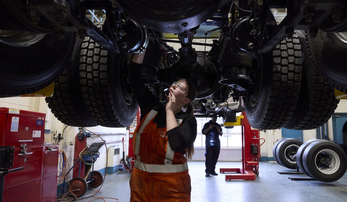 A mechanic wearing safety glasses and an orange work overall inspects the underside of a large truck lifted on a hydraulic hoist in a workshop, with tools and equipment visible in the background.