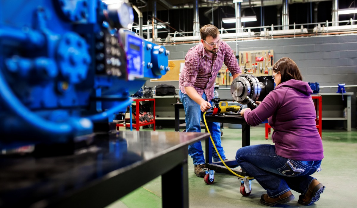 Two people wearing safety glasses work together in a workshop, inspecting and adjusting a large mechanical component on a table, with industrial equipment and tools visible in the background.