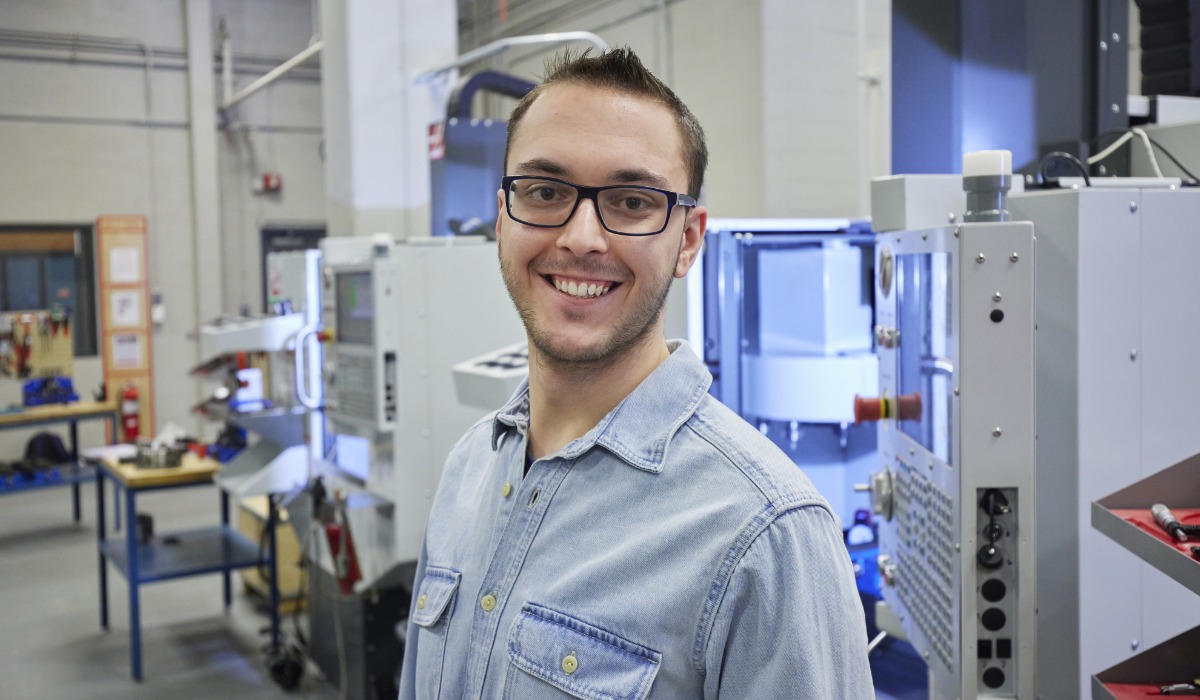 “A smiling person wearing glasses and a denim shirt stands in a workshop with machining equipment and tools in the background.