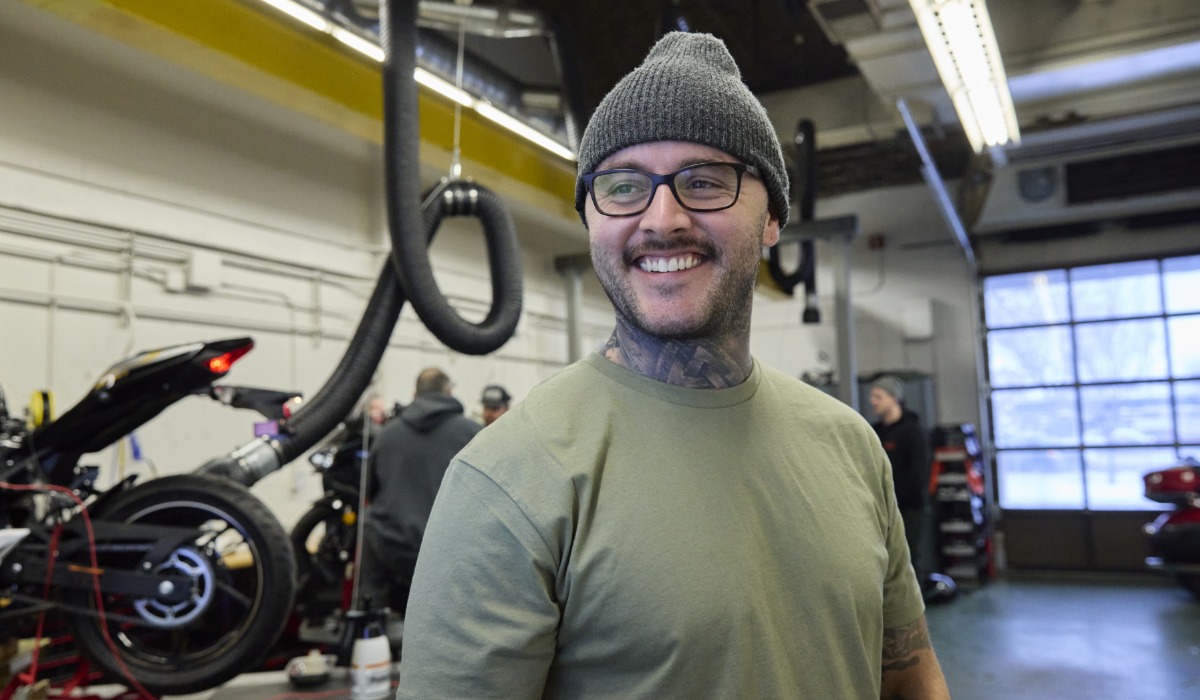 A smiling person wearing glasses, a beanie, and a green shirt stands in a motorcycle workshop with bikes, tools, and other mechanics working in the background.