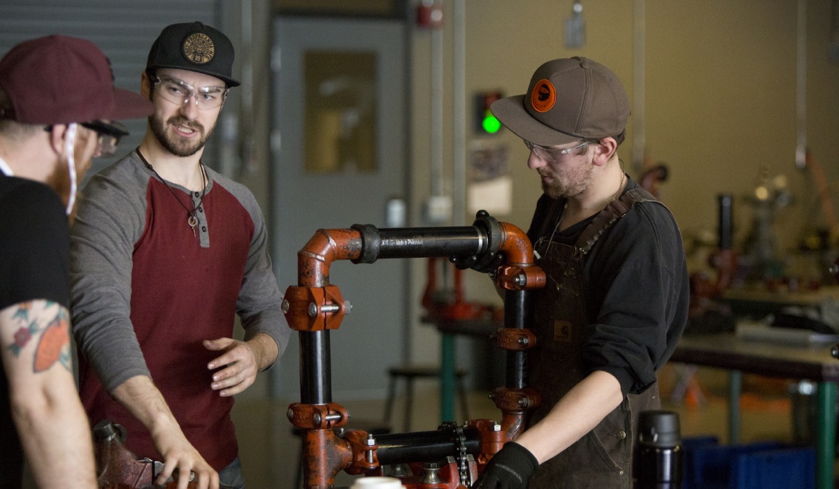 “Three people wearing safety glasses and work clothes assemble a metal pipe structure in a workshop, discussing and adjusting the components together.