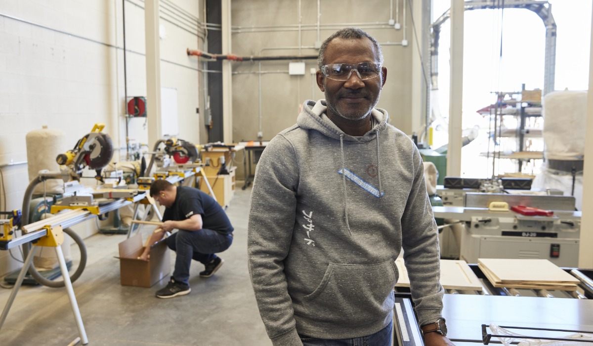 A person wearing safety glasses and a gray hoodie stands in a workshop with woodworking and mechanical equipment, while another person works with materials in the background.