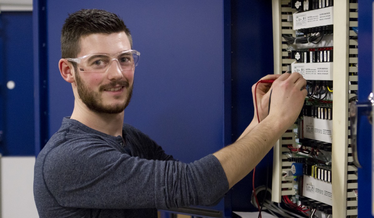 A person wearing safety glasses works on electrical wiring inside a control panel, using testing equipment and smiling toward the camera.