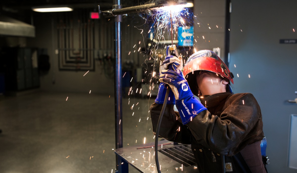 “A welder wearing protective gloves, a helmet, and safety gear works on a vertical metal pipe, with bright sparks flying in a workshop setting.