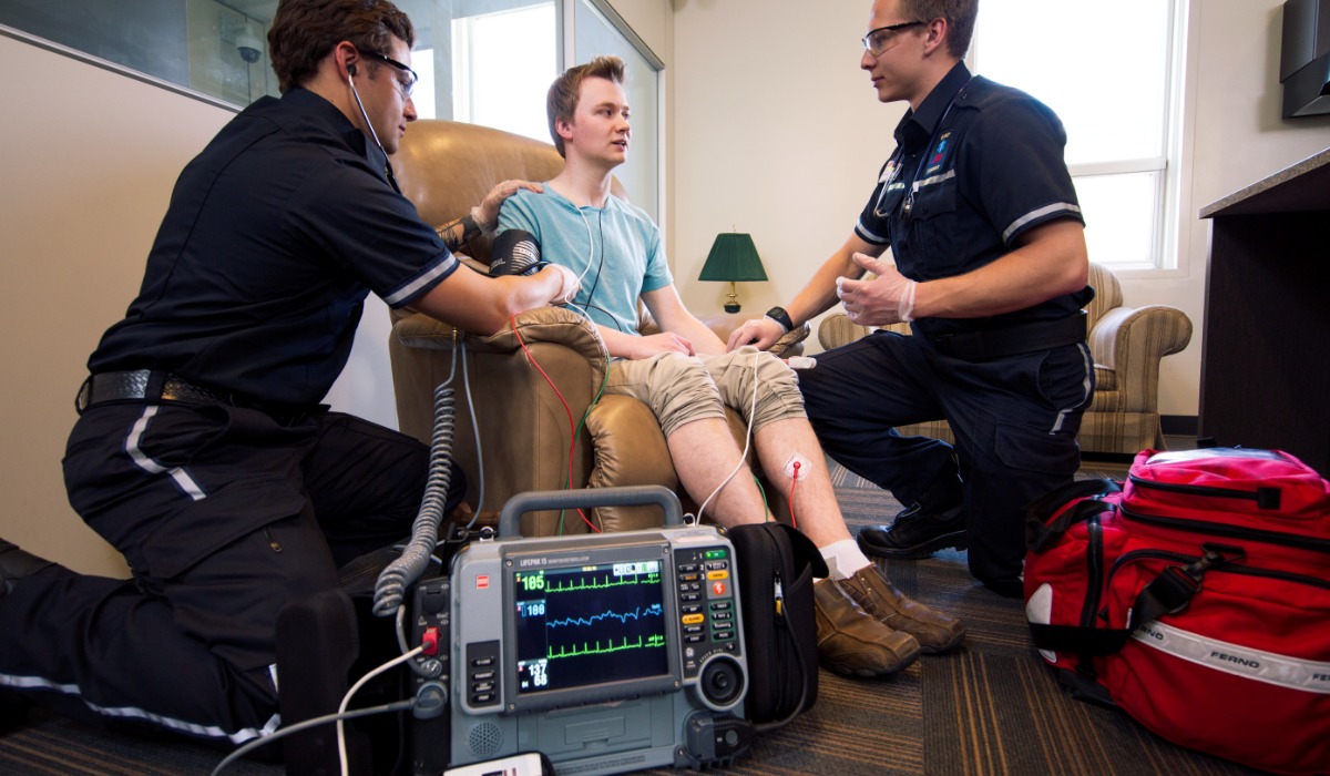 Medical personnel attending to a patient seated in a chair, with monitoring equipment visible in the foreground.