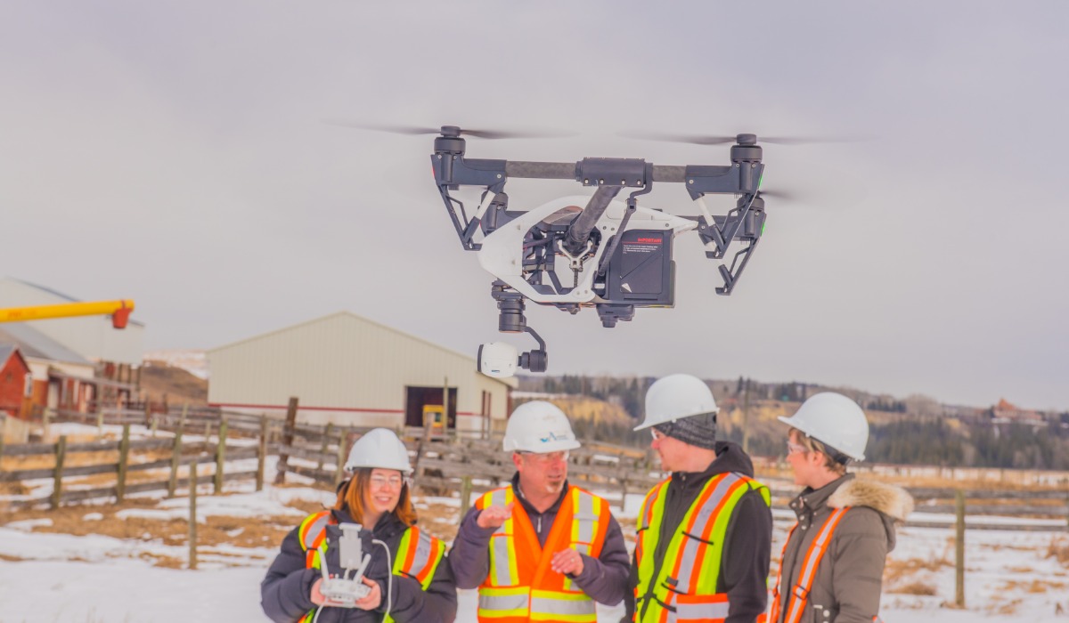 A drone is flying above a group of four individuals in safety vests and helmets, gathered in a rural setting with buildings and fences in the background.