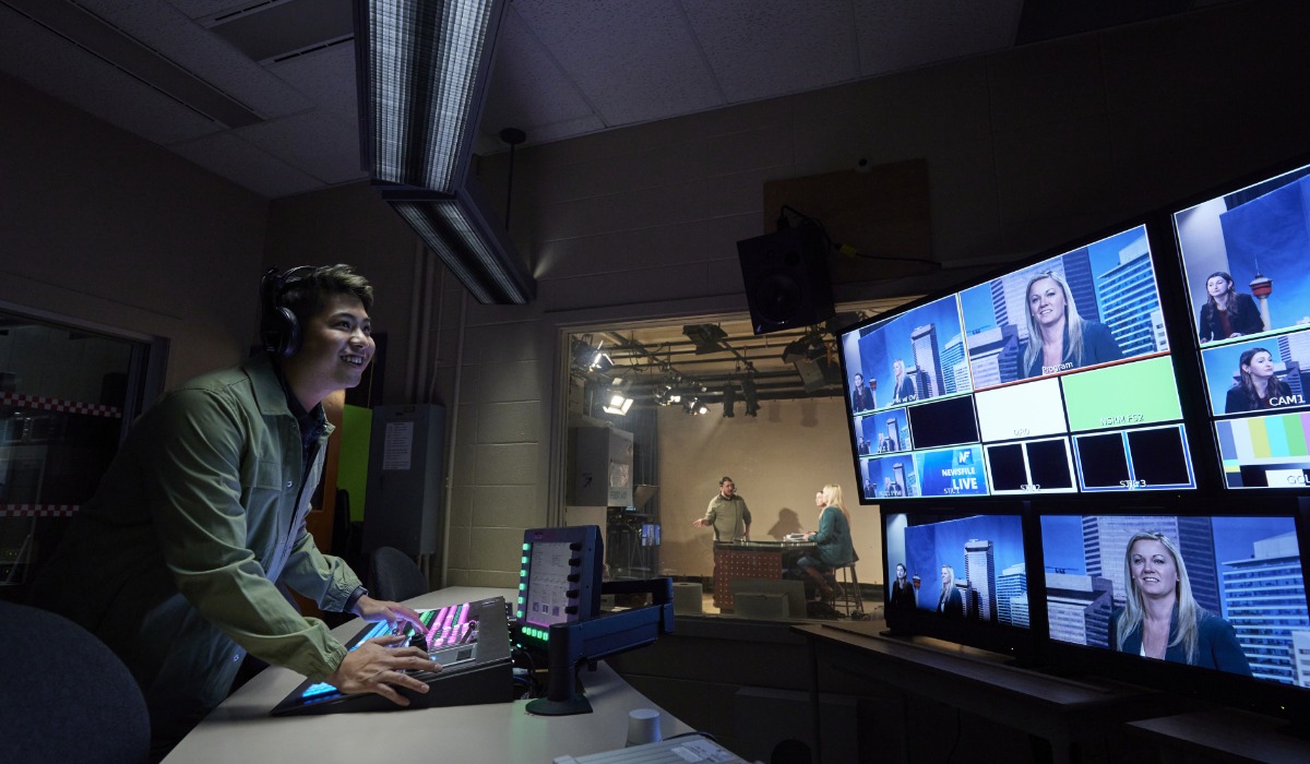 A television production control room featuring a smiling technician operating a control panel while monitors display various camera feeds and on-air talent.