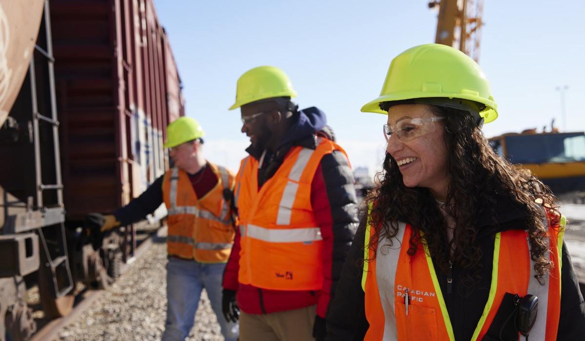 Three railway workers wearing hard hats and safety vests smile while inspecting a freight train.