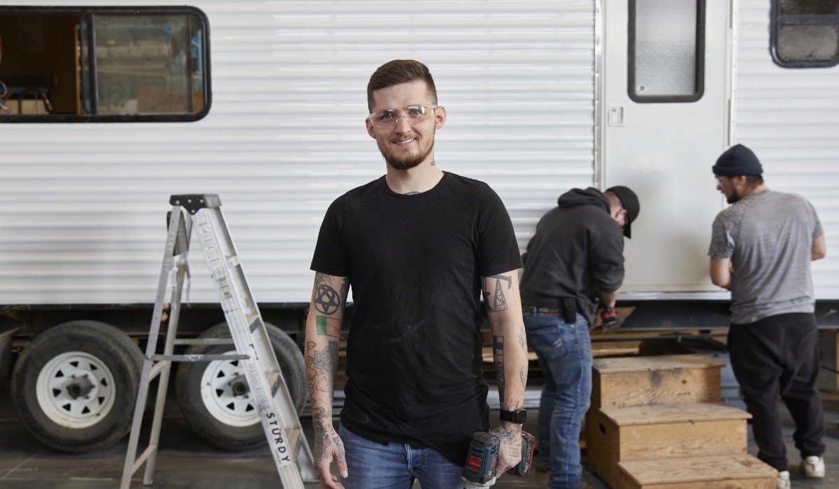 An apprentice wearing safety glasses and holding a power drill stands smiling in front of a trailer under repair, while two others work on steps leading to its door.