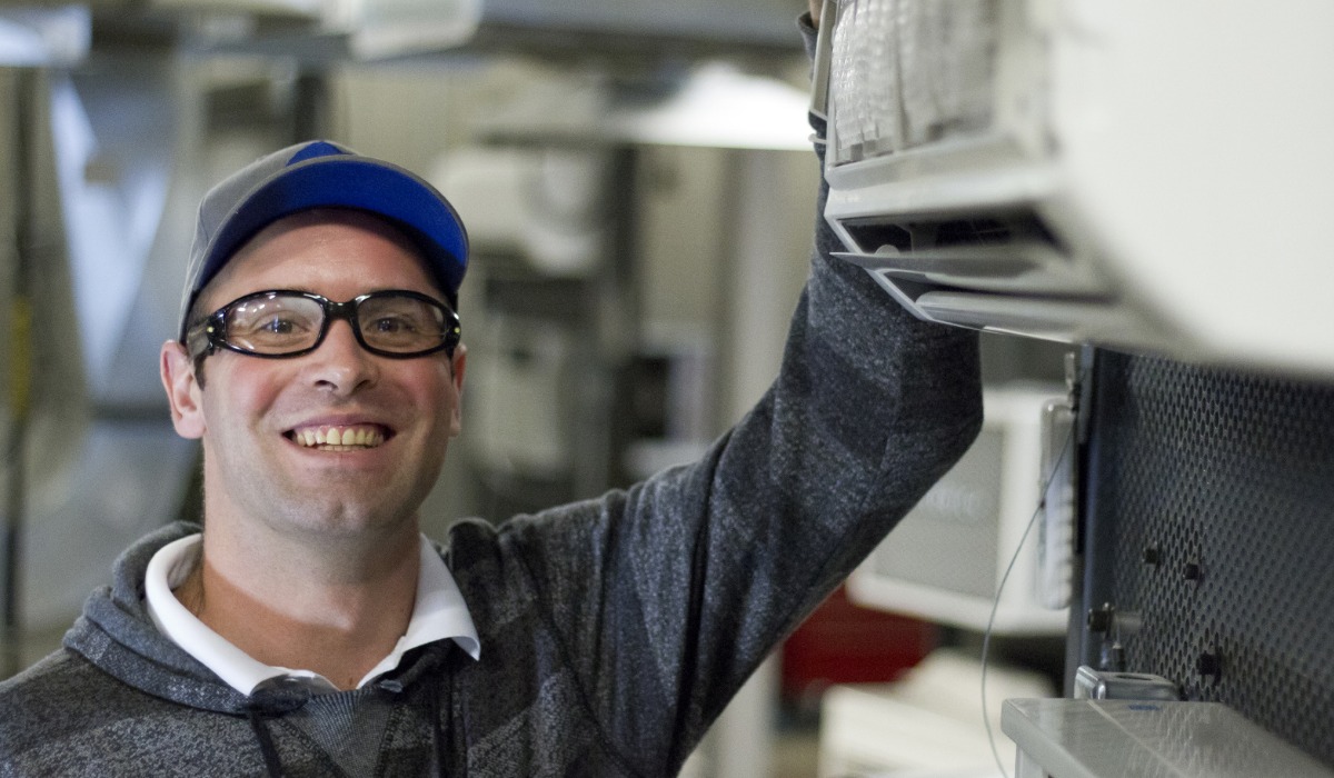 A smiling apprentice wearing safety glasses and a cap stands next to an air conditioning unit, appearing to inspect or service the equipment.