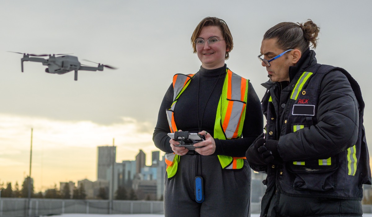 Two individuals wearing safety vests are operating a drone against a city skyline backdrop.
