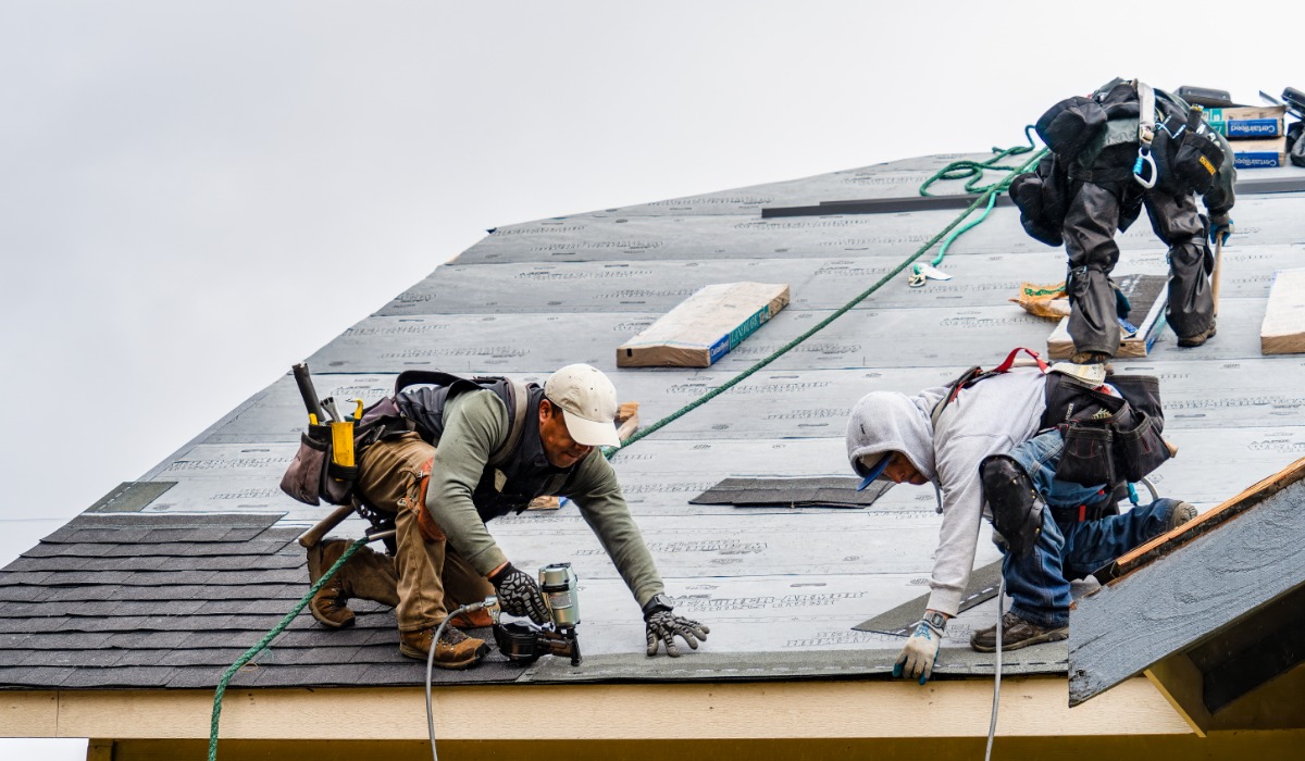 Three roofers wearing safety harnesses and work gear install shingles on a sloped roof, using tools and materials spread across the surface.