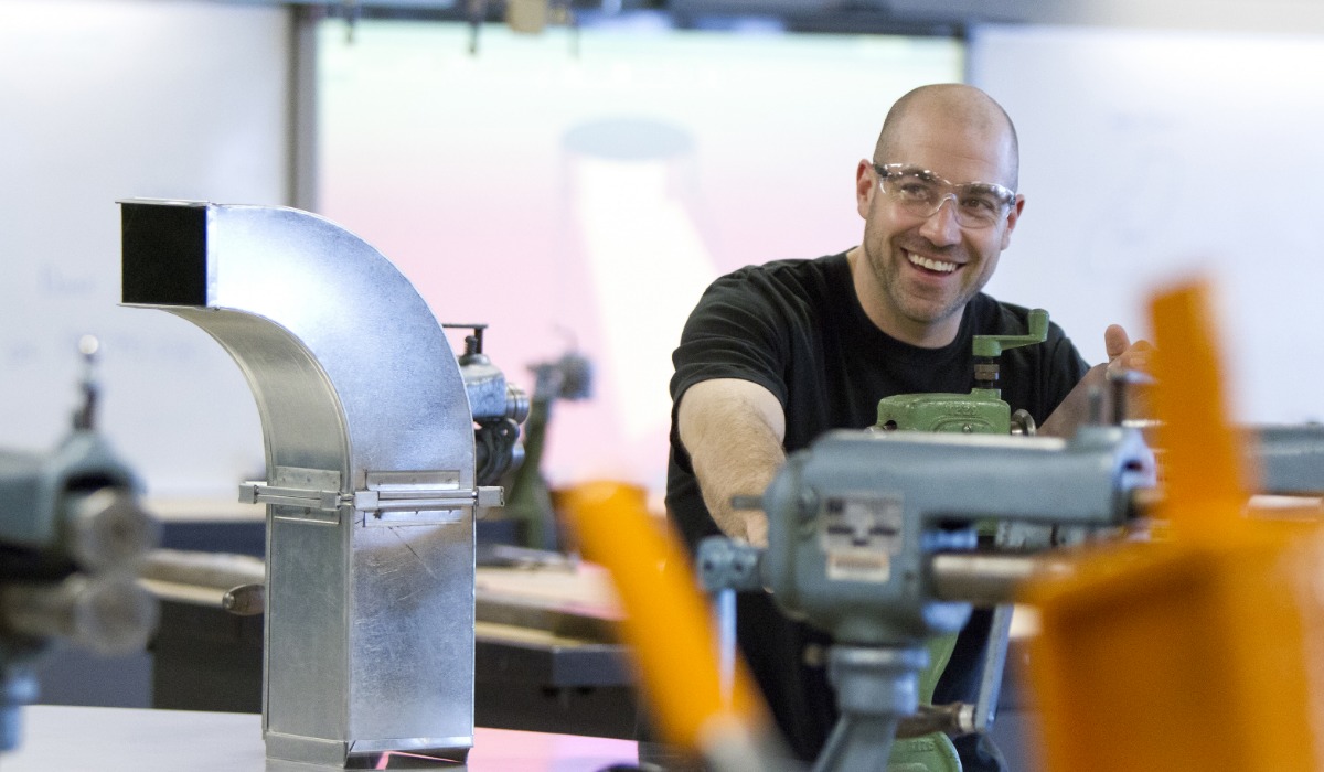 A smiling sheet metal worker wearing safety glasses operates machinery in a workshop, with metal duct components and tools on the workbench.