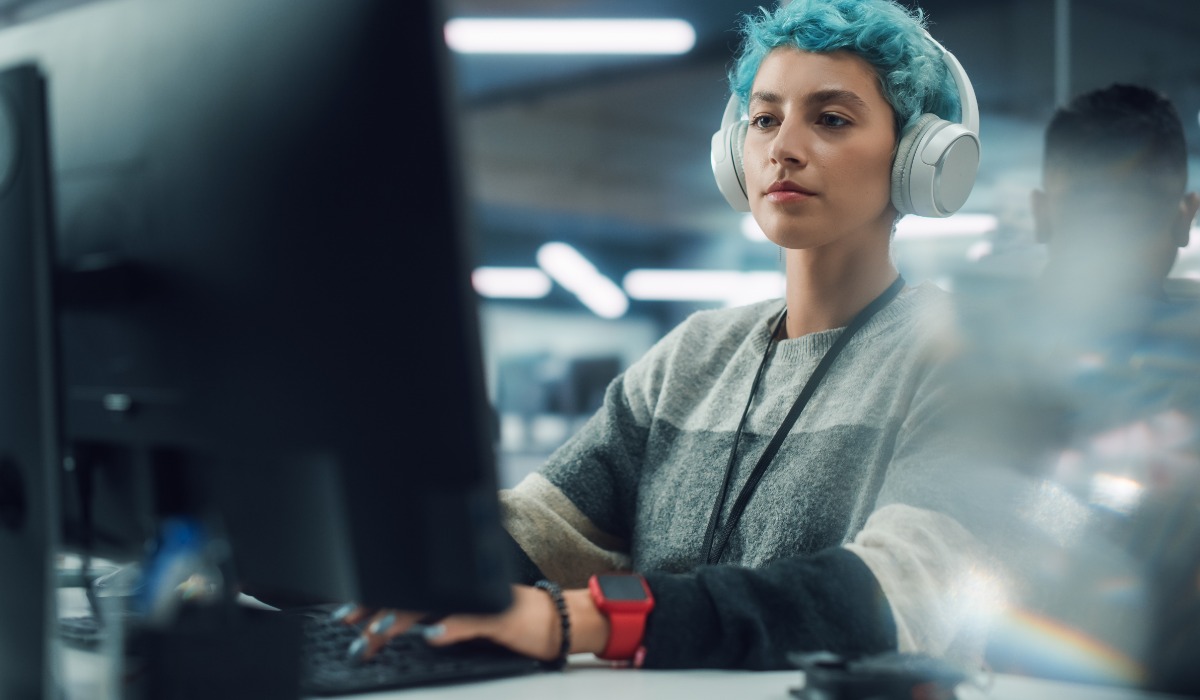 A focused woman with blue hair wearing headphones, working intently on a computer in a modern office setting.