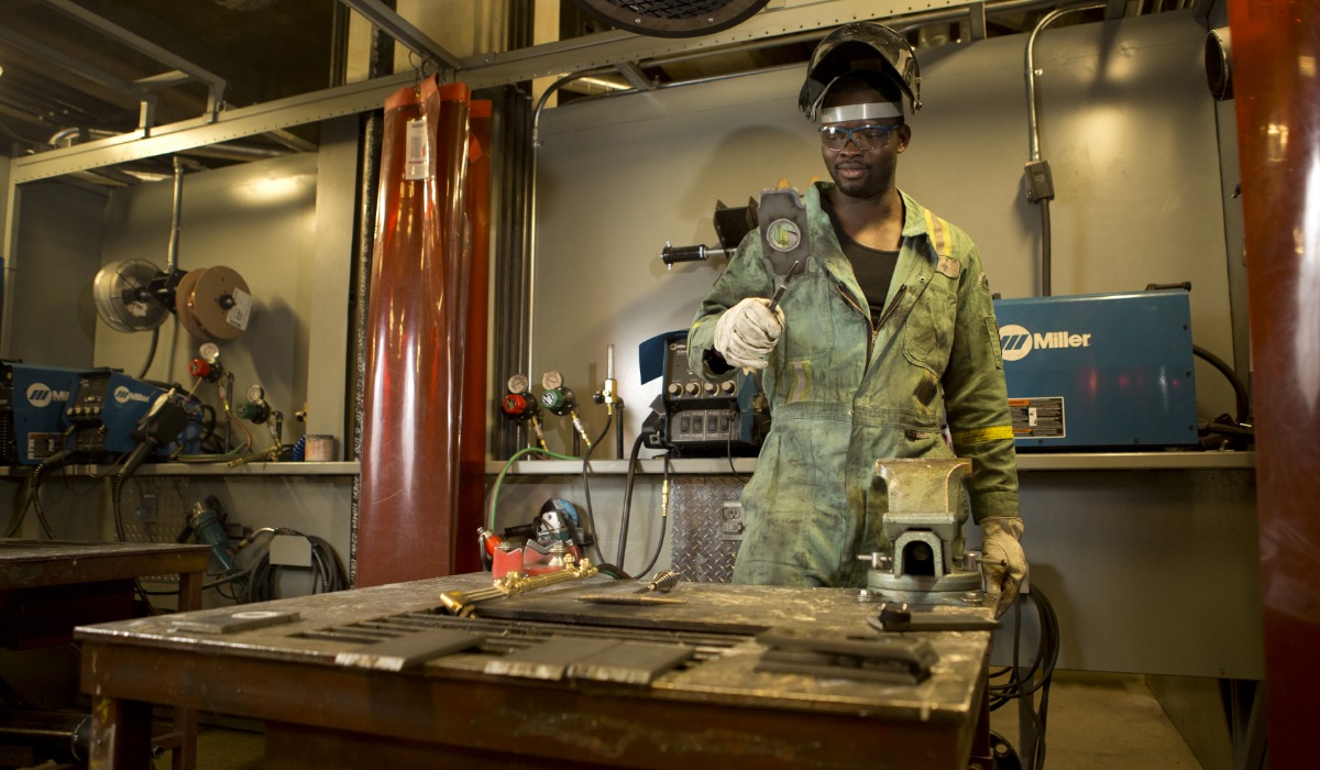 An apprentice wearing safety gear and a welding helmet stands in a metal fabrication workshop holding a tool, surrounded by welding equipment and workbenches