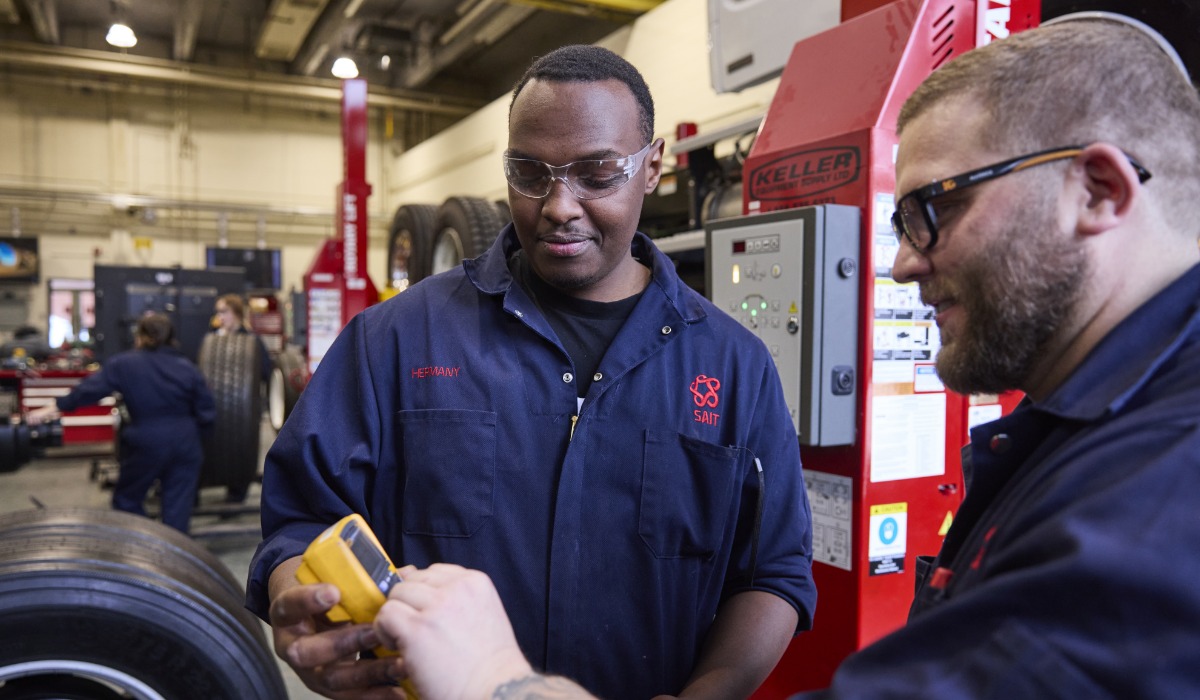 Two mechanics in a workshop collaborate on a diagnostic tool while working on a vehicle tire.