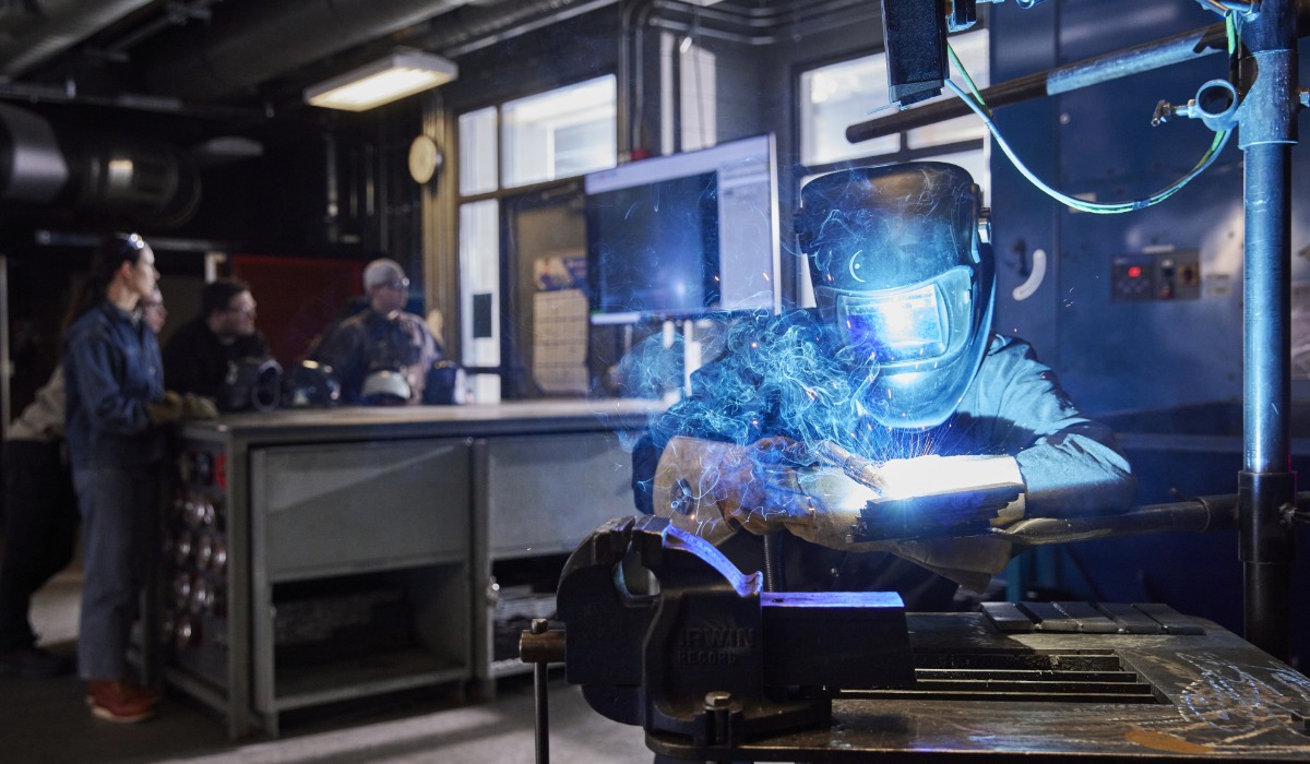 A welder wearing protective gear and a helmet works on a metal piece with bright sparks flying, while a group of people observe from a distance in the workshop.