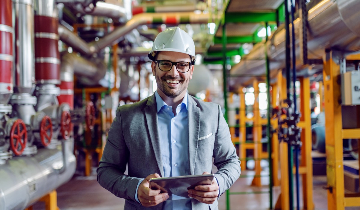 A smiling engineer wearing a suit, safety glasses, and a white hard hat holds a tablet while standing in an industrial facility with metal pipes, valves, and machinery in the background.