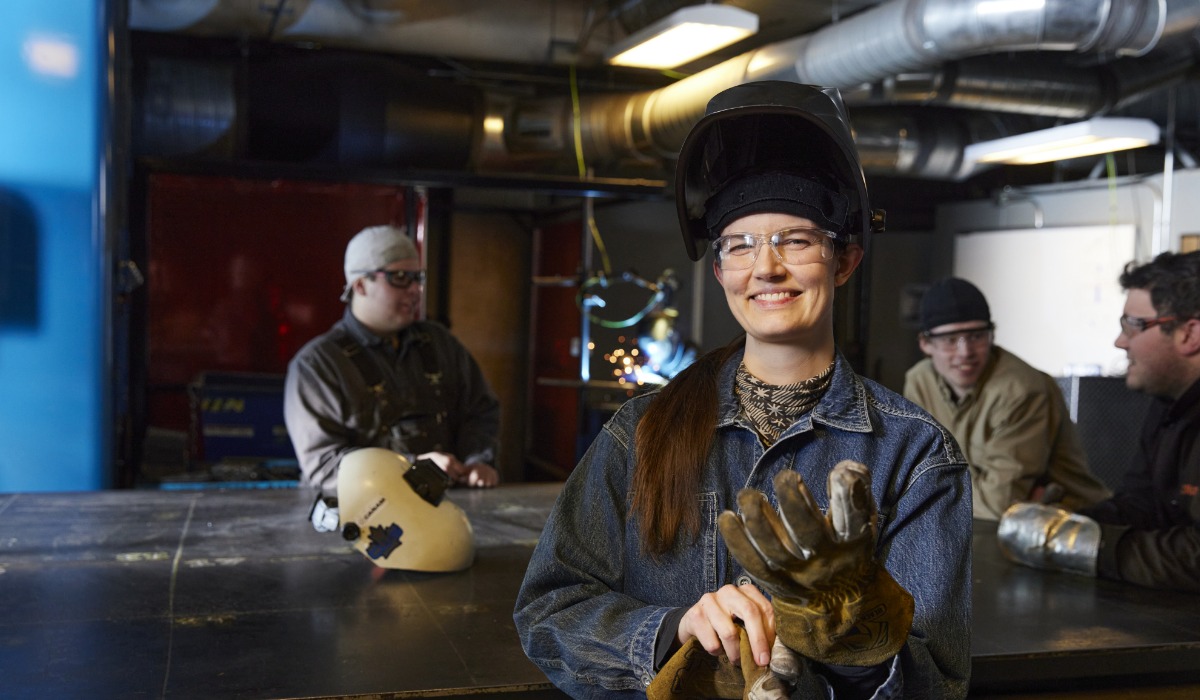 Smiling woman in welding gear stands in a workshop, showcasing confidence and readiness for the task. Other individuals are engaged in various activities in the background.