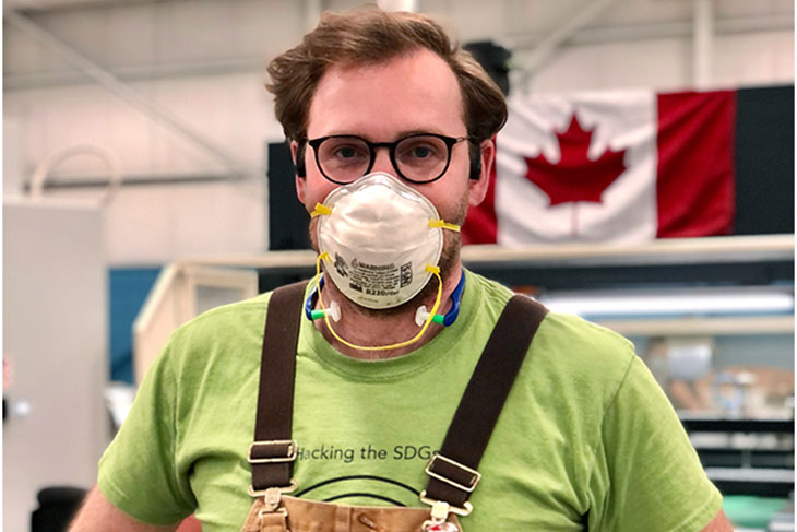 Doug Braden standing in the middle of factory floor with hands on hips, wearing khaki overalls, green tshirt, face mask and gloves