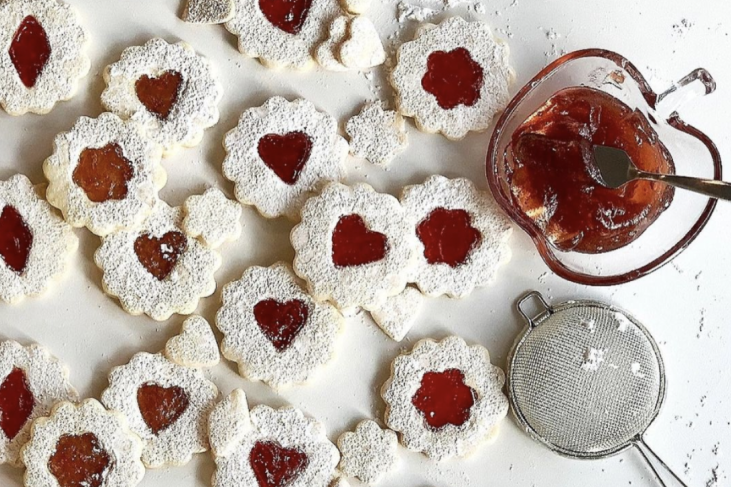 Short dough cookies spread on a white counter, filled with raspberry jam in heart and flower shaped cutouts. There's a jar of jelly and a sifter for icing sugar next to the cookies.