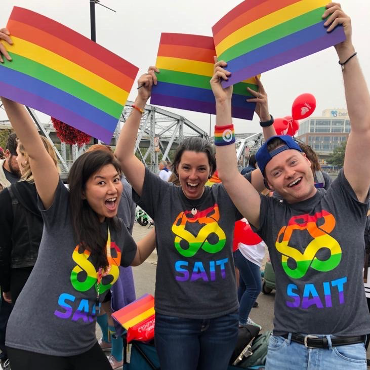 Three people wearing shirts with the SAIT rainbow logo smiling and holding up Pride flags 