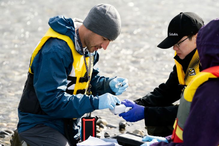 Students in life jackets analyzing water samples next to a river