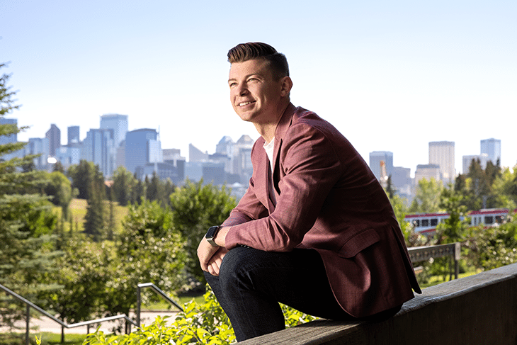 Kyle Bukauskas sitting on concrete wall ledge with city skyline behind him