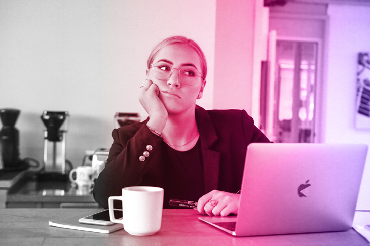  A person wearing glasses and a dark blazer looks uninterested at work and rests her chin on her hand while sitting at a desk in front of a laptop, cup of coffee, phone and notebook. The image is black and white on the left side and gradually fades into a bright pink and purple colour on the right side.