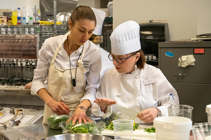 Two chefs in a kitchen are looking at greens during meal preparation.