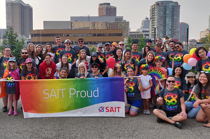 A group of people wearing SAIT pride shirts pose with a rainbow banner that says 'SAIT Proud'