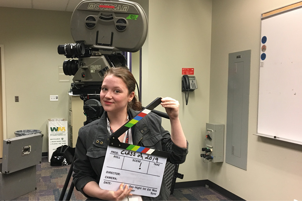 Dailies Editor and SAIT grad Jessica Rovansek holds a film clapperboard in front of a movie camera.