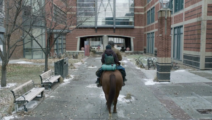 A view from behind Joel and Ellie as they ride a horse through SAIT’s campus. Trees and a red-bricked building are on either side of them.