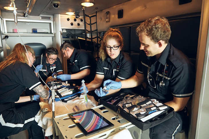 Five students in the back of an ambulance wearing paramedic uniforms hold medical equipment and work together.