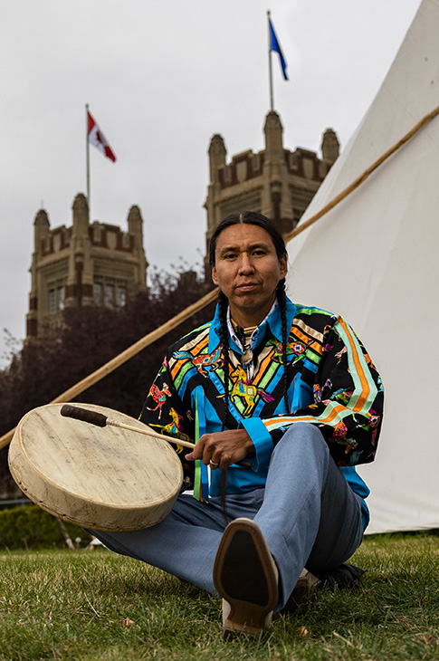 Indigenous man with drum sitting on grass in front of a tipi
