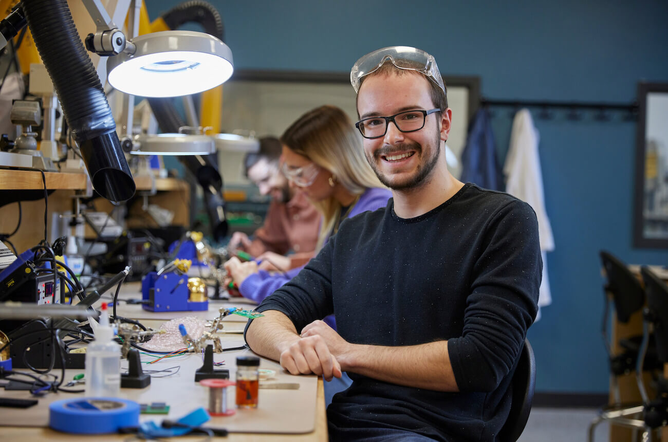 An electrician apprentice wearing safety glasses smiles for the camera while sitting at his work bench with other students.